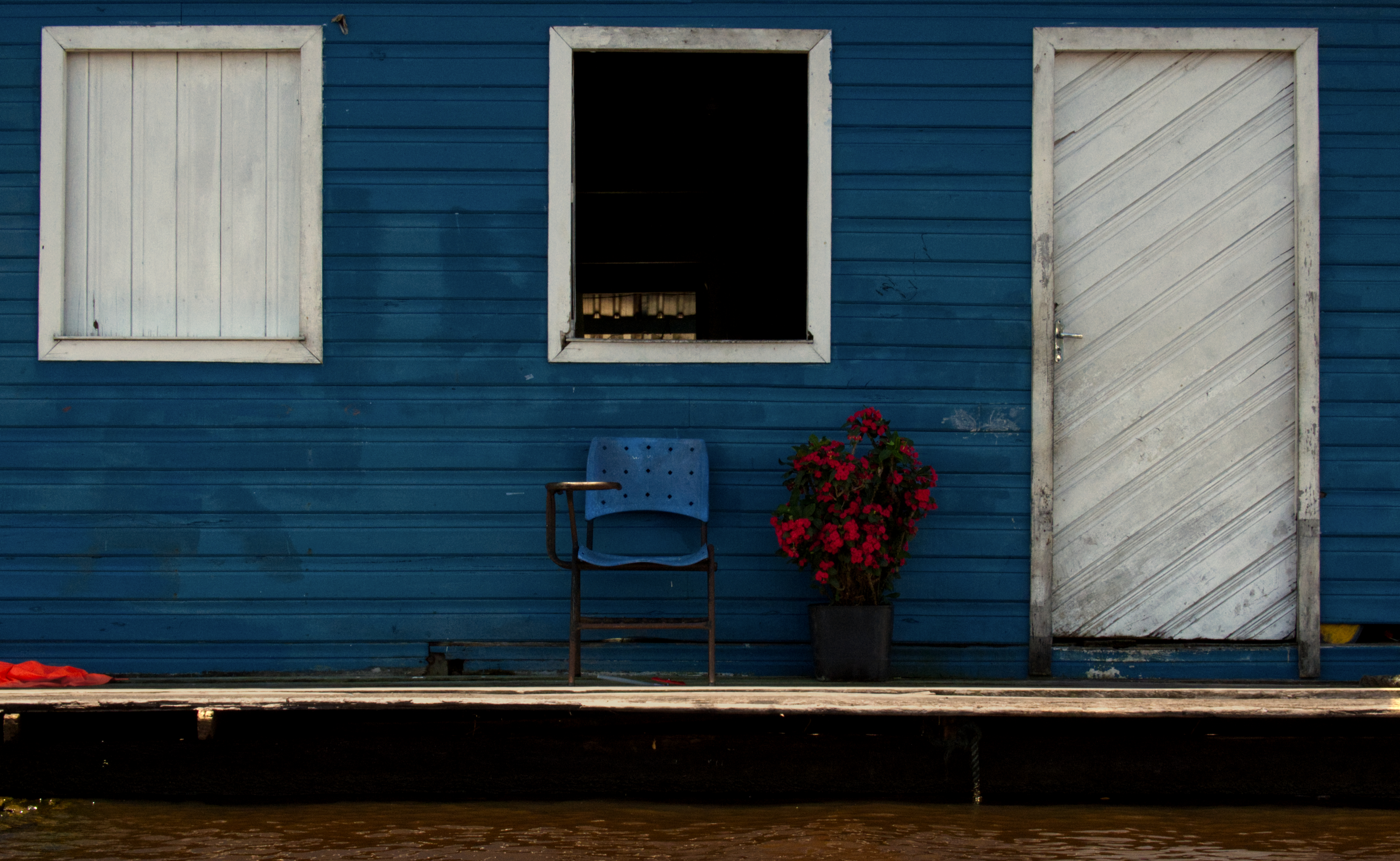 Homes line the riverbank, or during high water, sit atop it entirely. They are oriented toward water as the primary route of movement. Entry points, platforms, and openings face the river, reflecting its role in daily transport and access. Many are painted in deep, saturated colors: the blue of the houses along this stretch is striking against the pale sediment-heavy water.